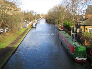 The image depicts a serene canal scene, featuring a green and red boat, a grassy bank, and a row of houses, set against a bright and sunny day.