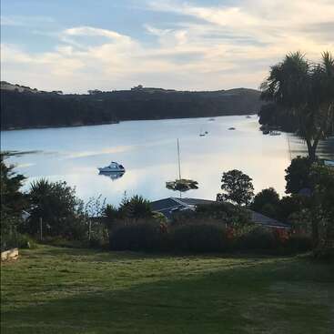 L'image représente une scène de lac sereine avec des bateaux, entourée de verdure et d'arbres luxuriants, sur fond de ciel nuageux. Une maison est visible au premier plan.