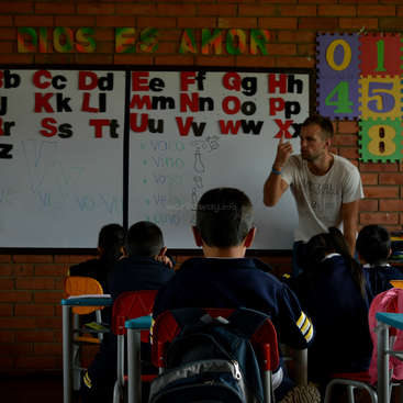 The image depicts a teacher instructing a group of students in a classroom, with a whiteboard displaying the alphabet and numbers, and a brick wall in the background.