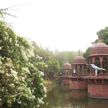 A serene scene features ornate red stone pavilions over a calm waterway. Two people practice yoga poses near lush greenery and white flowering bushes. Peaceful ambiance.