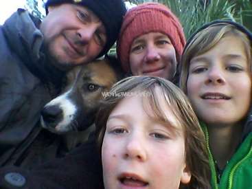 Das Bild zeigt eine vierköpfige Familie, die für ein Selfie posiert, mit einem Hund im Hintergrund. Die Familie ist in warme Winterkleidung gekleidet.