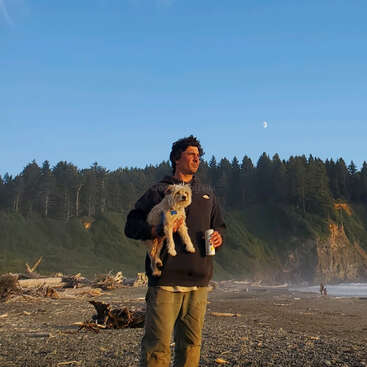 A man stands on a beach, holding a small dog and a drink, with a forested hillside and moon in the background, set against a clear blue sky.