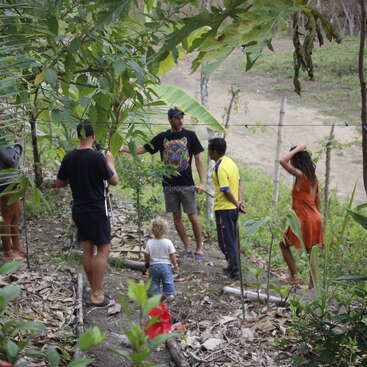 The image depicts a group of people, including children and adults, standing in a garden or yard, surrounded by lush greenery and trees, engaged in conversation.