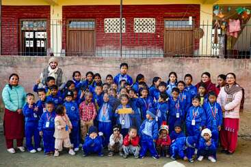 The image depicts a group of approximately 40 children, accompanied by three adults, posing for a photo in front of a building. The children are dressed in blue tracksuits.