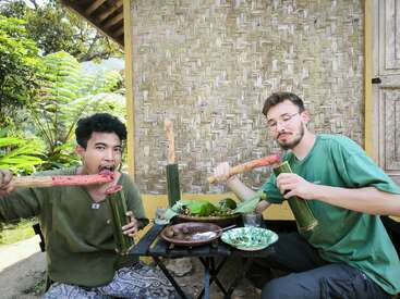 Two men sit at a table, eating from bamboo sticks, with a woven wall and lush greenery in the background, evoking a relaxed outdoor setting.