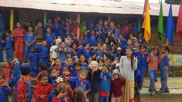 The image depicts a group of children in blue and red tracksuits posing for a photo with an adult, standing in front of a building with a corrugated roof.