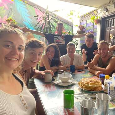 The image depicts a group of seven people seated at a table, surrounded by a vibrant atmosphere with plants and colorful walls, enjoying a meal together.
