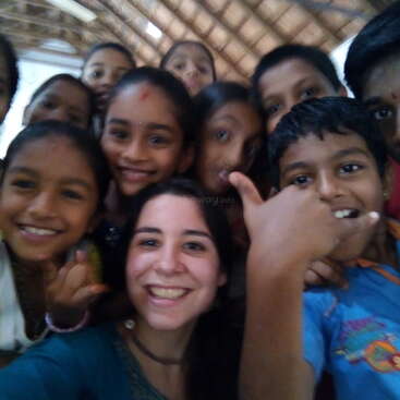 The image shows a group of children posing for a selfie with a woman, likely a teacher or volunteer, in a classroom setting with a tiled roof.