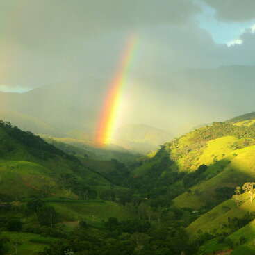 The image depicts a vibrant rainbow stretching across a lush green valley, set against a backdrop of rolling hills and mountains under a cloudy sky.