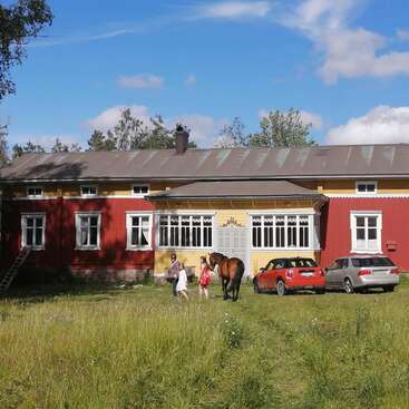 The image depicts a large red house with a brown roof, featuring white-framed windows, surrounded by a lush green field, with a horse and two people standing in front.