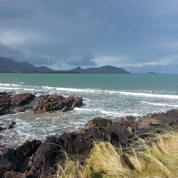 The image depicts a rocky coastline with a body of water and mountains in the background, under a cloudy sky.