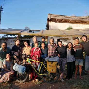 The image depicts a group of people standing in front of a building, with a yellow bicycle in the foreground, set against a clear blue sky.