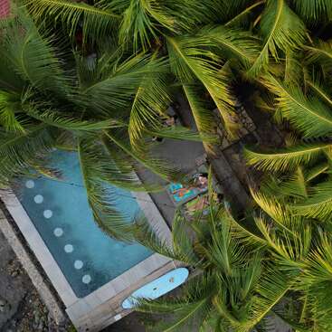 This aerial photo shows a serene pool surrounded by lush palm trees, a surfboard, stone pathways, and a person relaxing on a colorful towel in paradise.
