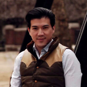 The image depicts a man with dark hair, wearing a white shirt and brown vest, standing in front of a blurred background, possibly outdoors.