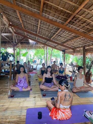 The image depicts a serene yoga class in a bamboo hut, with a group of individuals seated on mats, surrounded by lush greenery and natural light filtering through the open roof.