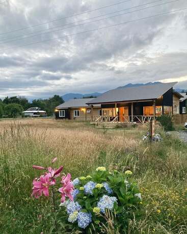 The image depicts a serene rural scene featuring a house with a porch, surrounded by a field of tall grass and wildflowers, set against a cloudy sky.