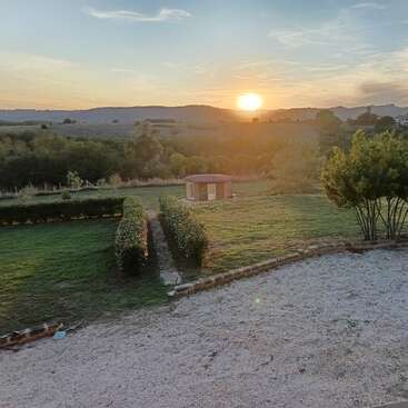 A beautiful countryside scene at sunset, featuring a green lawn, neatly trimmed hedges, a small shed, trees, gravel area, and rolling hills in the background.