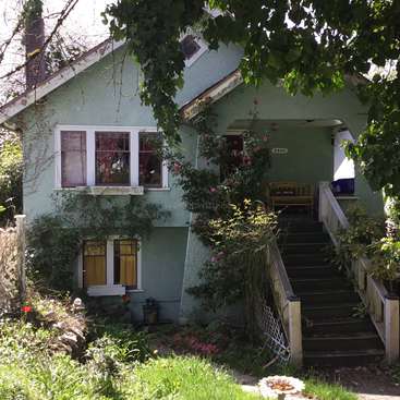 The image depicts a charming light green house with a covered porch, surrounded by lush greenery and vibrant pink flowers, exuding a sense of serenity and tranquility.