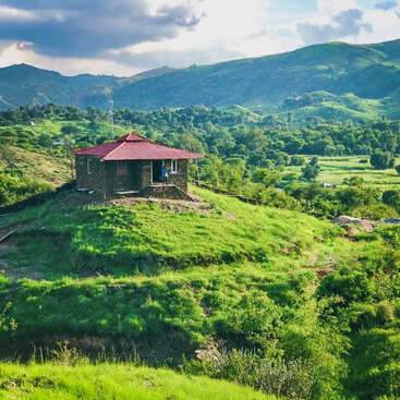 The image depicts a serene stone house with a red roof, nestled on a hillside amidst lush greenery, with a person standing in the doorway, set against a mountainous backdrop.