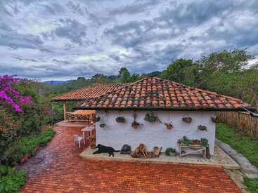 The image depicts a serene white house with a tiled roof, surrounded by lush greenery and vibrant flowers, set against a cloudy sky.