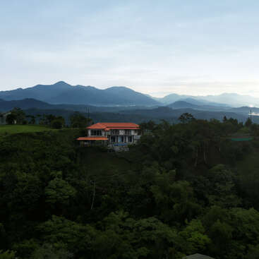 The image depicts a serene scene of a house with a red roof nestled among lush green trees, set against the majestic backdrop of distant mountains under a cloudy sky.