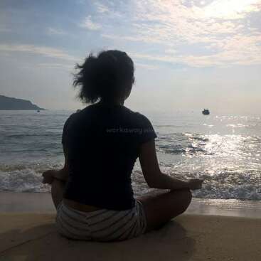 A person sits cross-legged on a sandy beach facing the ocean, meditating peacefully at sunrise or sunset, with calm waves and a distant boat.