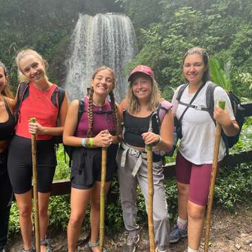 The image shows five women standing in front of a waterfall, each holding a hiking stick and wearing backpacks and casual clothing, posing for a photo.
