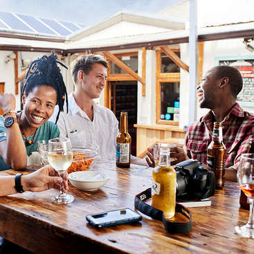 The image depicts three individuals seated at a wooden table, surrounded by drinks and snacks, with a camera and phone nearby, set against a backdrop of a building with a white exterior and wooden accents.