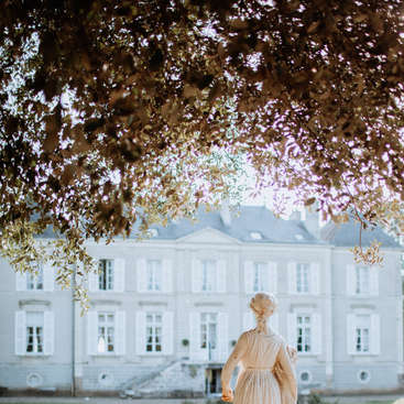 L'image représente une scène sereine d'une statue de femme en robe blanche, sur fond d'un grand bâtiment de couleur claire avec de nombreuses fenêtres.