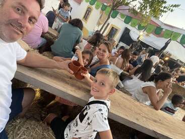The image depicts a family of four sitting at a long wooden table, toasting with clay cups, surrounded by a festive atmosphere with hay bales and green flags.