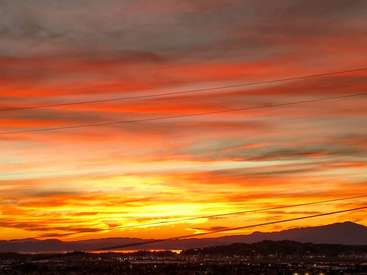 The image depicts a vibrant sunset over a cityscape, with a mountain range in the background and power lines stretching across the sky.