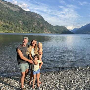 The image depicts a family of four posing on a rocky beach, with a serene lake and majestic mountains serving as a picturesque backdrop under a blue sky with white clouds.