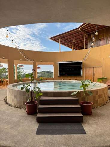 An inviting circular pool with wooden steps, surrounded by potted plants. String lights hang above, with a bar and seating area set against warm, tan walls.