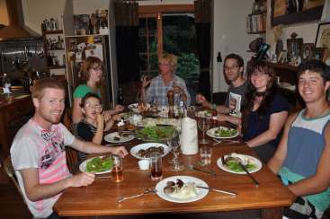 The image depicts a family of eight seated around a wooden table, enjoying a meal together in a cozy home setting. The atmosphere is warm and inviting.