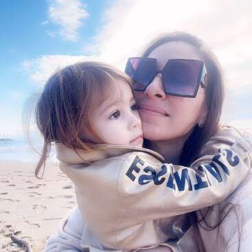 The image shows a woman and a child on a beach, with the woman wearing sunglasses and holding the child in her arms. The child is wearing a beige jacket.
