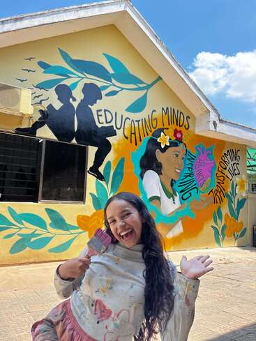 A cheerful woman stands in front of a colorful mural she painted, featuring positive messages about education, inspiration, and transformation, with vibrant flowers and silhouettes.
