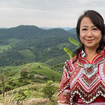 The image depicts a woman standing in front of a lush green mountainous landscape, wearing a vibrant red and white embroidered top with intricate patterns and designs.
