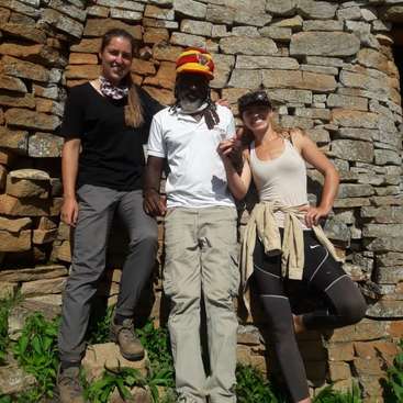 The image depicts three individuals standing in front of a stone wall, posing for a photo, with two women on either side of a man, all dressed in casual outdoor attire.