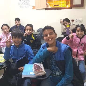 A group of smiling children sit in a classroom, some holding up peace signs and thumbs up, appearing happy and cheerful, with notebooks on their desks.