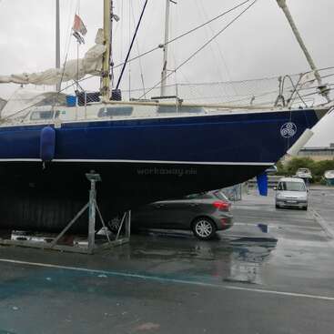 The image shows a dark blue sailboat on a boat lift in a wet parking lot, with a gray car parked underneath it.