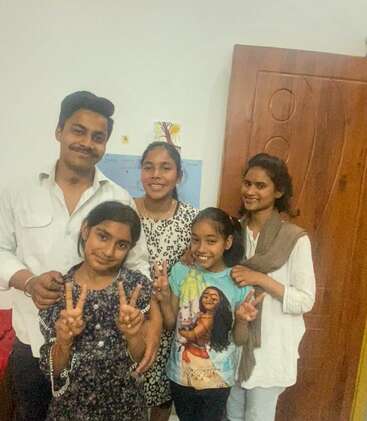 L'image montre une famille de cinq personnes posant pour une photo, le père se tenant derrière la mère et les trois filles, qui font toutes des signes de paix.