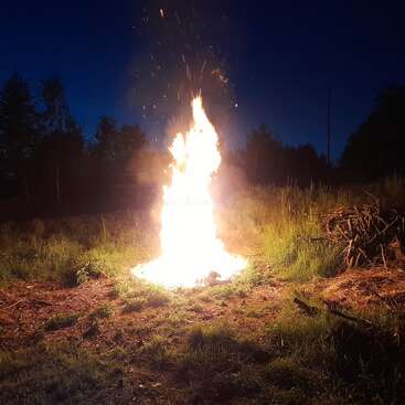 A large, bright bonfire blazes intensely in a grassy outdoor area at night, surrounded by darkness, trees, and a pile of branches nearby. Sparks rise.