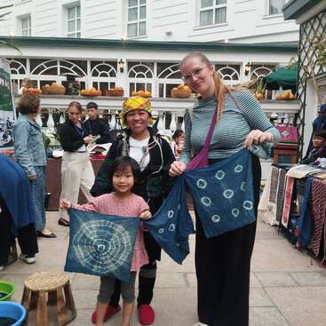 The image depicts two women and a child standing together, proudly holding up indigo-dyed textiles, with a bustling outdoor market scene in the background.