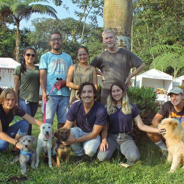 The image depicts a group of eight people posing with four dogs on a grassy area, surrounded by trees and white tents in the background.
