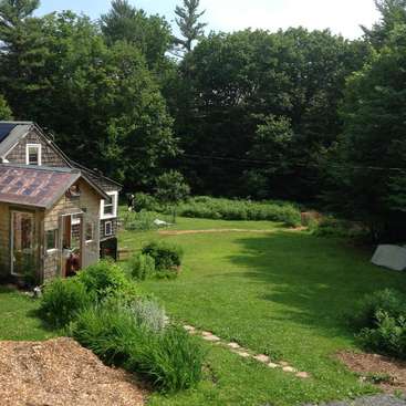 The image depicts a charming small house with a brown roof and beige siding, nestled among lush greenery and trees, exuding a sense of serenity and tranquility.