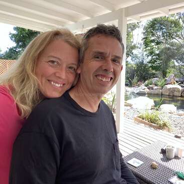 The image depicts a man and woman posing for a photo on a porch, with a serene pond and garden visible in the background.