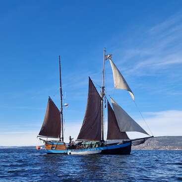 The image depicts a sailboat with three sails on a large body of water, set against a blue sky with wispy clouds and a distant shoreline.