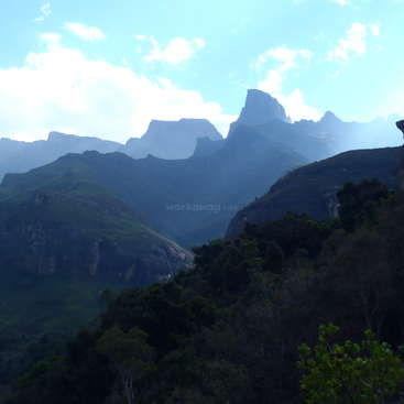 The image depicts a mountain range with lush greenery, set against a blue sky with white clouds, evoking a sense of natural beauty and serenity.