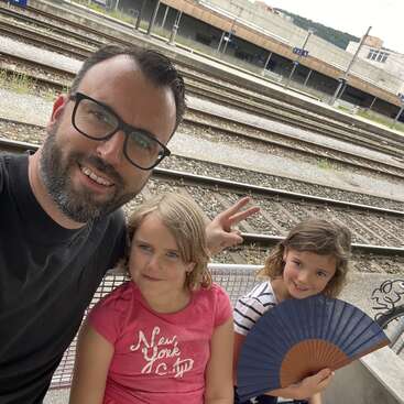 The image shows a man and two young girls posing for a selfie in front of train tracks, with the man on the left and the girls on the right.