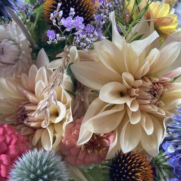 The image presents a vibrant bouquet of flowers, featuring a mix of white, pink, yellow, and purple blooms, with a prominent cream-colored dahlia taking center stage.